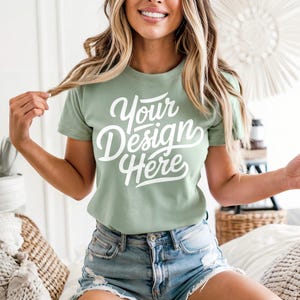 May include: Sage green t-shirt with the text "Your Design Here" in a white script font. The shirt is short-sleeved and paired with denim shorts. The model is smiling and holding a strand of hair.