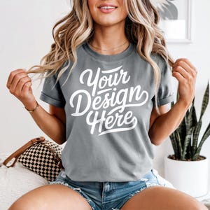 May include: Gray t-shirt with the text "Your Design Here" in a white, cursive font. The shirt has short sleeves and a crew neck. The model is wearing blue denim shorts.