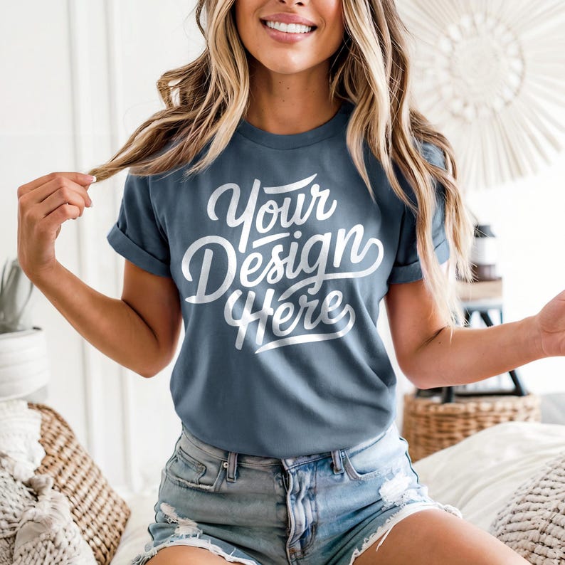 May include: A blue t-shirt with the text "Your Design Here" in a white script font. The shirt has rolled-up sleeves and is paired with denim shorts. The model is smiling and holding a strand of hair.