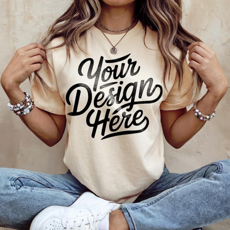 Pode incluir: Camiseta creme com o texto "Your Design Here" em preto. A camiseta &eacute; combinada com jeans azuis e t&ecirc;nis brancos. A modelo usa pulseiras e colares.