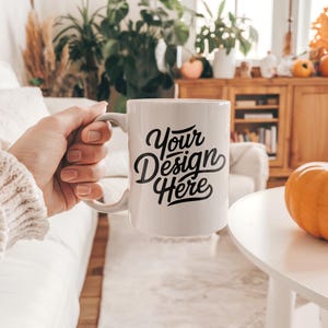 Puede incluir: Taza de cerámica blanca con un diseño negro que dice "Your Design Here". La taza está siendo sostenida por una mano en un suéter blanco.