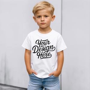 May include: A young child wearing a white t-shirt with the text "Your Design Here" in black script. The child is also wearing blue jeans and has their hands in their pockets. The background is a plain, light grey wall.