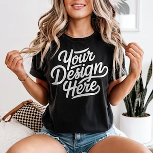 May include: Black t-shirt with the text "Your Design Here" in white script. The shirt has rolled-up sleeves and is paired with denim shorts. The model is wearing a gold necklace and bracelet.