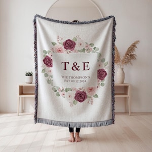 May include: Cream-colored woven throw blanket with a floral wreath design. The wreath features burgundy and pink roses, with the initials "T & E" and the date "09.12.2024" in the center. The blanket has a fringed edge.