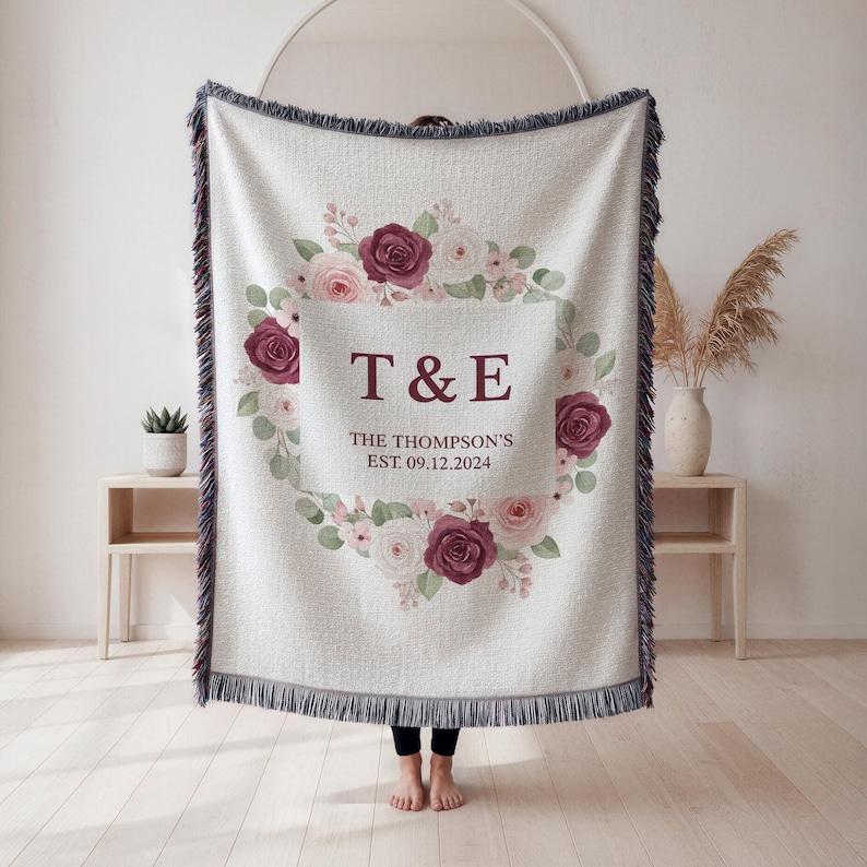 May include: Cream-colored woven throw blanket with a floral wreath design. The wreath features burgundy and pink roses, with the initials "T & E" and the date "09.12.2024" in the center. The blanket has a fringed edge.