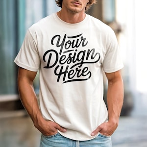 May include: Cream-colored t-shirt with the text "Your Design Here" in a black, cursive font. The shirt has short sleeves and a crew neck. The person is wearing blue jeans and has their hands in their pockets.
