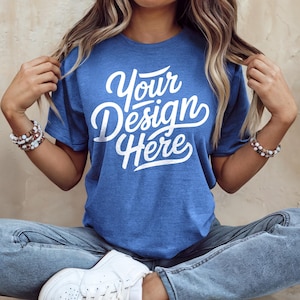 以下が含まれることがあります： 白い文字で「Your Design Here」と書かれた青いTシャツを着た女性。彼女は足を組んで座っており、青いジーンズと白いスニーカーを履いています。