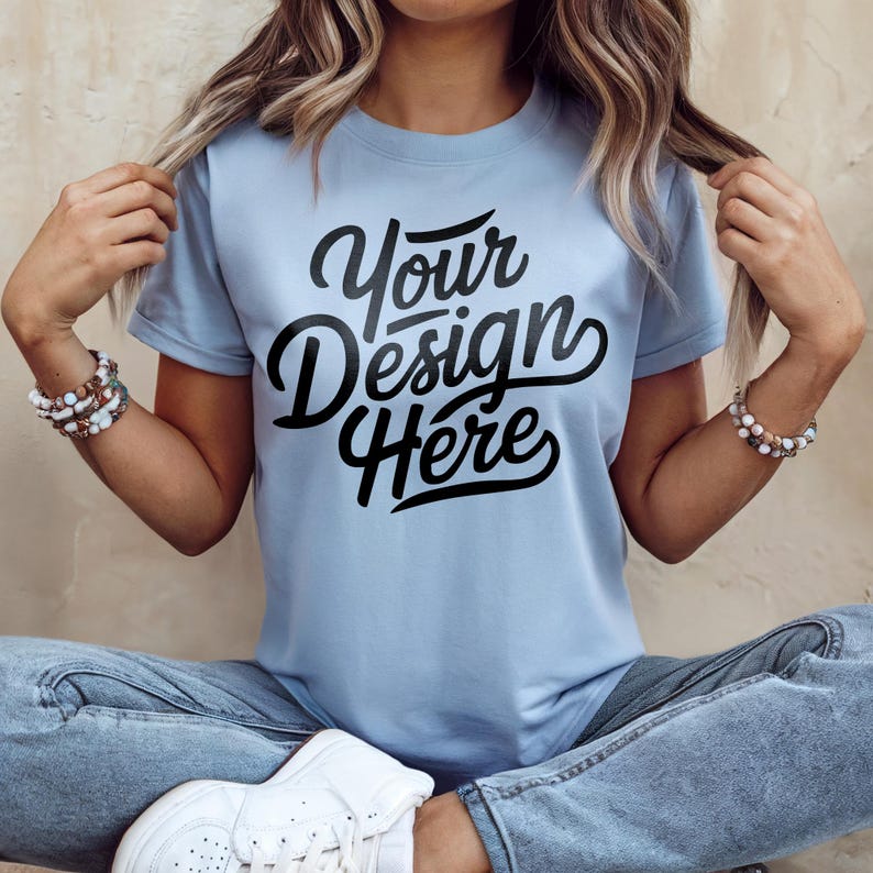 May include: A light blue t-shirt with the text "Your Design Here" in black script. The short-sleeved shirt is worn with blue jeans and white sneakers. The person is wearing bracelets.