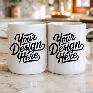 May include: Two white ceramic mugs with a curved handle. Each mug features the black text "Your Design Here" in a stylish script font. The mugs are set against a blurred background.