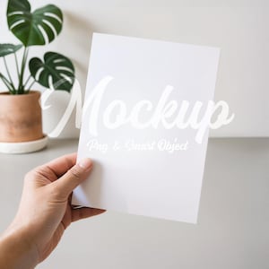 Può includere: Un biglietto bianco vuoto tenuto in mano, con la parola "Mockup" in un carattere corsivo. Sotto la parola "Mockup" c'è il testo "Png & Smart Object". Una pianta in vaso è sullo sfondo.