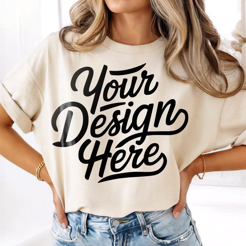 Puede incluir: Camiseta color crema con el texto "Your Design Here" en escritura negra. La camiseta tiene las mangas enrolladas y se combina con vaqueros azules. Se ve una pulsera dorada en la mu&ntilde;eca.