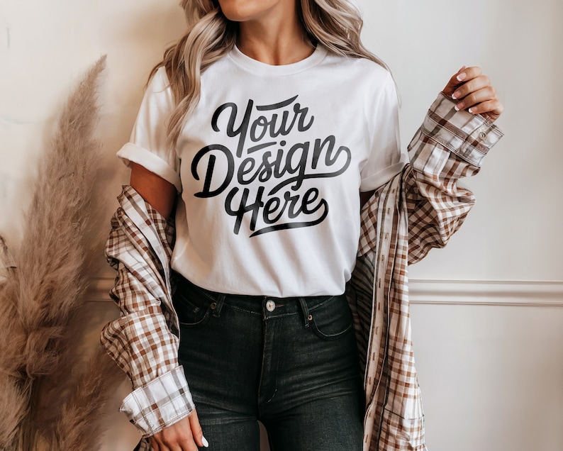 May include: White t-shirt with the text "Your Design Here" in black script font. The t-shirt is being worn by a person with long blonde hair and a brown and white plaid shirt.