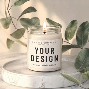 May include: A clear glass candle jar with a white label that says "CANDLE COMPANY TYPE OF CANDLE YOUR DESIGN Net Wt 8oz | Burn Time of 40 hours". The candle is lit and the flame is visible.