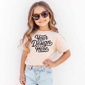 May include: A beige t-shirt with the text "Your Design Here" in black script. The t-shirt is worn by a young person wearing sunglasses and light blue denim jeans. The background is a plain white wall.