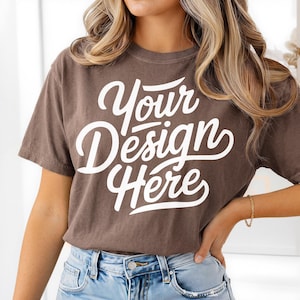 Może przedstawiać: Brązowa koszulka z napisem "Your Design Here" w białym skrypcie. Koszulka z krótkim rękawem jest noszona z niebieskimi dżinsami. Modelka ma na sobie złotą bransoletkę.