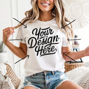 May include: White short-sleeved t-shirt with the text "Your Design Here" in a black script font. The shirt is being worn with blue denim shorts. The model is smiling and holding her hair.
