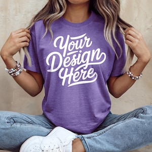May include: A purple t-shirt with the text "Your Design Here" in a white script font. The shirt is short-sleeved and has a crew neck. The model is wearing blue jeans and white sneakers.