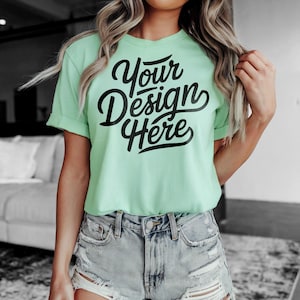 May include: A light green t-shirt with the text "Your Design Here" in black lettering. The shirt is being worn by a person with long blonde hair and is paired with light blue denim shorts.