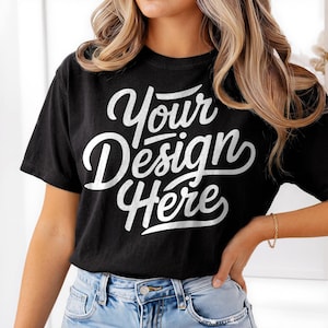 Könnte beinhalten: Schwarzes T-Shirt mit dem weißen Schriftzug "Your Design Here" in kursiver Schrift. Das kurzärmelige Shirt scheint aus einem weichen Material zu sein. Das Model trägt eine blaue Jeans und ein goldenes Armband.