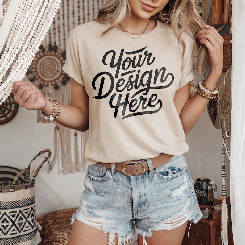 May include: A beige t-shirt with the text "Your Design Here" in black script font. The shirt is being worn by a person with long blonde hair and is paired with denim shorts and a brown belt.