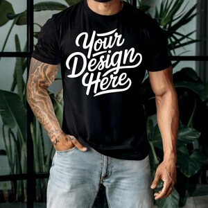 K&ouml;nnte beinhalten: Ein schwarzes T-Shirt mit dem Text "Your Design Here" in wei&szlig;en Buchstaben. Das Shirt wird von einer Person mit T&auml;towierungen auf den Armen getragen.