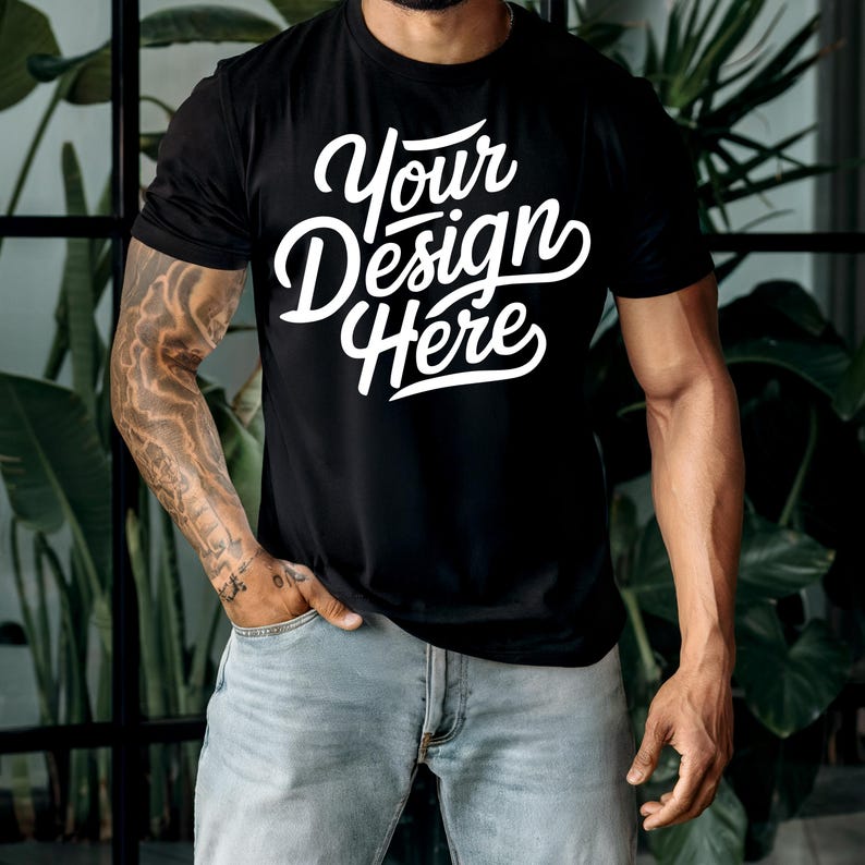 K&ouml;nnte beinhalten: Ein schwarzes T-Shirt mit dem Text "Your Design Here" in wei&szlig;en Buchstaben. Das Shirt wird von einer Person mit T&auml;towierungen auf den Armen getragen.