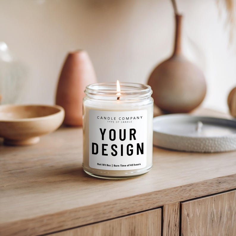 May include: A white candle with a black label that says "CANDLE COMPANY TYPE OF CANDLE YOUR DESIGN Net Wt 8oz | Burn Time of 40 hours". The candle is lit and sitting on a wooden surface.
