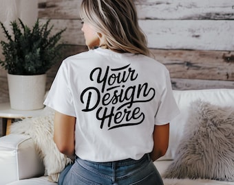 Könnte beinhalten: Ein weißes T-Shirt mit dem Text "Your Design Here" in schwarzer Schrift auf dem Rücken. Das T-Shirt wird von einer Person getragen. Der Hintergrund umfasst ein weißes Sofa, eine Pflanze und eine Holzwand.