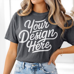 May include: Dark gray t-shirt with the text "Your Design Here" in white script. The shirt has a vintage, washed look and is paired with blue jeans. The model is wearing a gold bracelet.