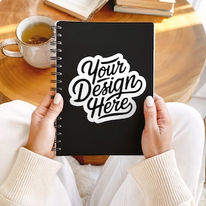 May include: A black spiral-bound notebook with the text "Your Design Here" in a white, stylized font. The notebook is held open, revealing the design. A cup of tea and books are in the background.
