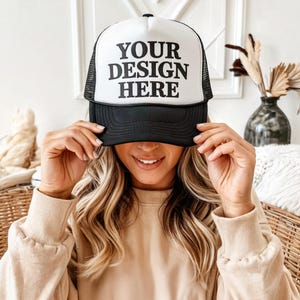 Puede incluir: Una gorra de camionero negra y blanca con una malla blanca en la parte posterior. La parte delantera de la gorra tiene el texto "YOUR DESIGN HERE" en negro.