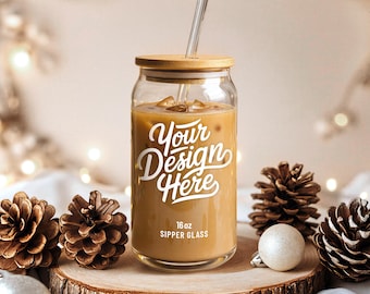 Puede incluir: Un vaso transparente para beber con tapa de bambú y pajita transparente, lleno de una bebida color café y cubitos de hielo. El vaso tiene el texto "Your Design Here" y "16 oz Sipper Glass". El vaso está sobre una superficie de madera.