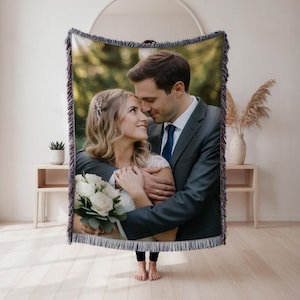 May include: Woven blanket featuring a wedding photo of a couple embracing. The blanket has a fringed edge and displays a color photograph of a bride and groom. The couple is dressed in formal attire, with the bride holding a bouquet of white flowers.