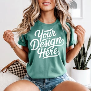May include: Teal-colored t-shirt with the text "Your Design Here" in white script. The shirt has rolled-up sleeves and is paired with denim shorts. A gold necklace and bracelet are visible.