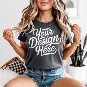 May include: Dark gray t-shirt with the text "Your Design Here" in white script. The shirt has short sleeves and is paired with denim shorts. The model is sitting on a bed with a patterned pillow.