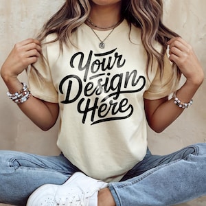 May include: Cream-colored t-shirt with the text "Your Design Here" in a bold, black script. The shirt is a relaxed fit, and the model is wearing blue jeans and white sneakers. The overall aesthetic is casual and comfortable.