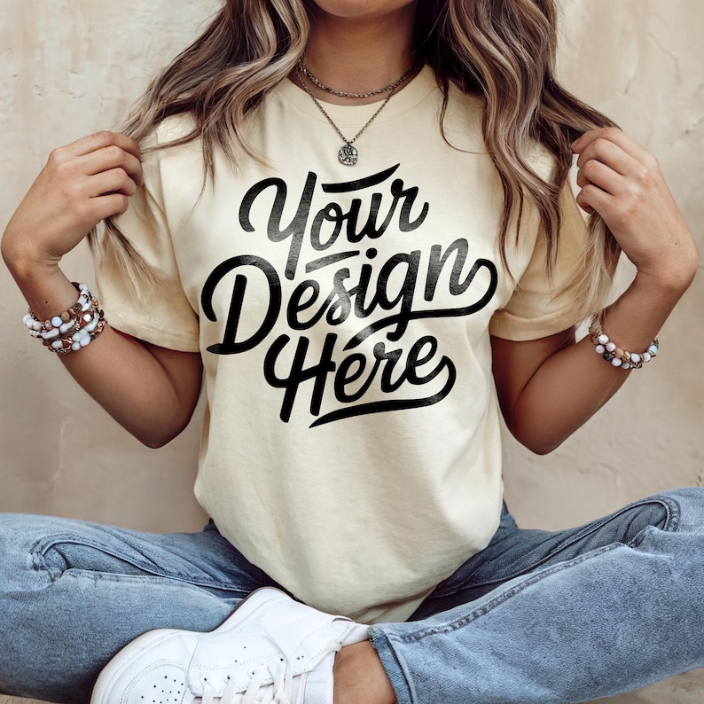 May include: Cream-colored t-shirt with the text "Your Design Here" in a bold, black script. The shirt is a relaxed fit, and the model is wearing blue jeans and white sneakers. The overall aesthetic is casual and comfortable.