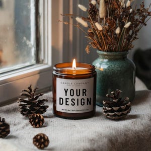 May include: A brown glass candle with a white label that says "CANDLE COMPANY TYPE OF CANDLE YOUR DESIGN Net Wt 8oz | Burn Time of 40 hours". The candle is lit and the flame is visible. The candle is sitting on a white surface with pine cones.