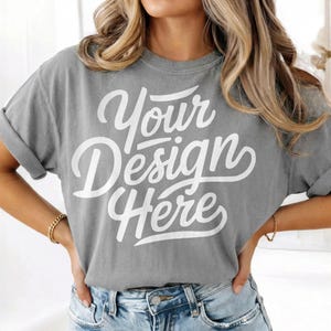 Peut inclure: Un t-shirt gris avec le texte "Your Design Here" en écriture blanche. Le t-shirt a des manches retroussées et est associé à un jean bleu. Le modèle porte un bracelet doré.