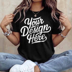 May include: Black t-shirt with the text "Your Design Here" in a white, cursive font. The shirt is being worn by a person wearing blue jeans and white sneakers. The person is also wearing bracelets and a necklace.