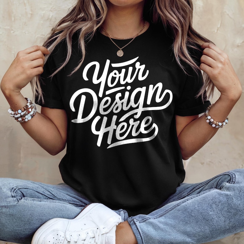 May include: Black t-shirt with the text "Your Design Here" in a white, cursive font. The shirt is being worn by a person wearing blue jeans and white sneakers. The person is also wearing bracelets and a necklace.