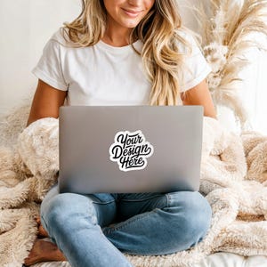 May include: A laptop with a sticker reading "Your Design Here" in black script with a white border. The laptop rests on a person's lap, who is wearing a white t-shirt and blue jeans.