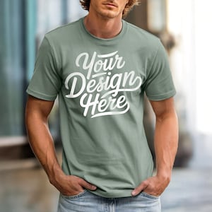 May include: Sage green t-shirt with the text "Your Design Here" in white script. The short-sleeved shirt is made of a soft material and is a blank canvas for custom designs. The model is wearing blue jeans.