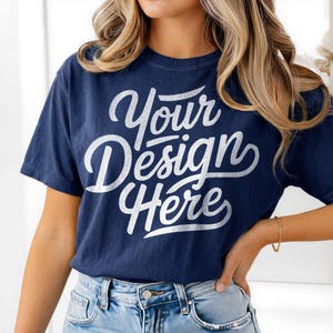 Pode incluir: Camiseta azul marinho com o texto branco "Your Design Here" em fonte cursiva. A camiseta é de manga curta e tem gola redonda. A modelo está usando jeans azuis e uma pulseira dourada.