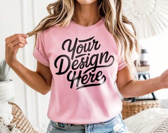 May include: A pale pink t-shirt with rolled sleeves. The shirt displays the text "Your Design Here" in a black, cursive font. The model is wearing denim shorts.