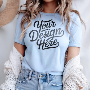 May include: A light blue t-shirt with the text "Your Design Here" in black lettering. The shirt is worn by a person with long blonde hair and is styled with a white cardigan and blue jeans.