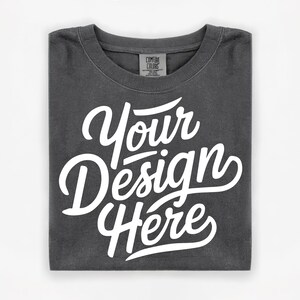 Peut inclure: T-shirt gris foncé plié, avec l'inscription blanche "Your Design Here" en écriture cursive. Le col affiche une étiquette Comfort Colors. Un vêtement vierge pour la personnalisation.