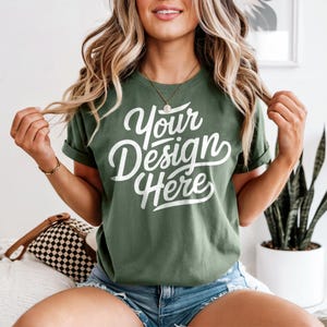 May include: Olive green t-shirt with rolled sleeves and the text "Your Design Here" in white script. The model is wearing denim shorts and a gold necklace. The shirt is a casual, everyday wear item.