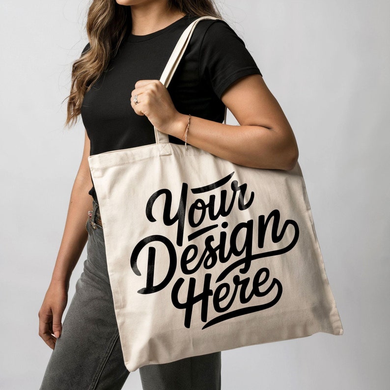 May include: A person is holding a white canvas tote bag with black text that says "Your Design Here".