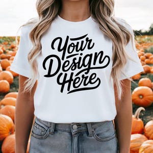 May include: White t-shirt with the text "Your Design Here" in black script font. The shirt is being worn by a person in a field of pumpkins.